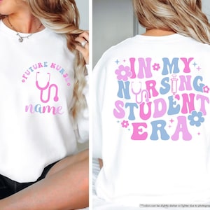 May include: White sweatshirt with a pink and blue design that says "Future Nurse" with a stethoscope and the word "name" on the front. The back of the sweatshirt says "In My Nursing Student Era" with a stethoscope and syringe in a pink and blue retro design.