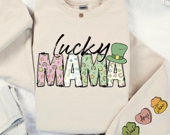 Op de afbeelding: Een crèmekleurige trui met de tekst "Lucky Mama" in zwart en groen. De tekst is omringd door een patroon van klavertjesvier, regenbogen en het woord "luck". De trui heeft een groene kabouterhoed in de rechterbovenhoek en drie patches op de rechtermouw met de namen "Noak", "Amy" en "Jane".