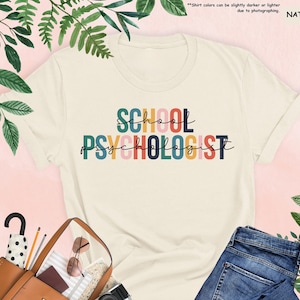 Op de afbeelding: Een wit T-shirt met de tekst "SCHOOL PSYCHOLOGIST" in regenboogkleuren. De tekst is in een gestileerd lettertype.
