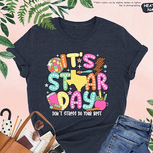 May include: A heather navy blue t-shirt with a colorful graphic design that says "It's Star Day" and "Don't Stress Do Your Best".