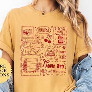 May include: Mustard-colored t-shirt with a red graphic featuring book-themed illustrations and text. The design includes phrases like "Grumpy x Sunshine," "Just One More Chapter," and "One Bed at the Inn." The shirt has a relaxed fit.
