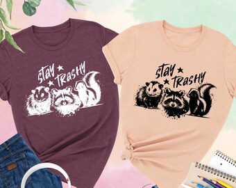 May include: Two t-shirts with a graphic of three animals, a raccoon, a skunk, and a sugar glider, with the text "stay trashy" printed on them. The shirts are in heather maroon and heather peach colors.