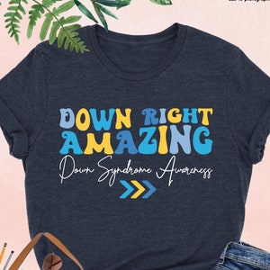 May include: A heather navy blue t-shirt with the text "DOWN RIGHT AMAZING" in colorful letters. The text "Down Syndrome Awareness" is written in a smaller font below the main text. Three yellow arrows point to the right.