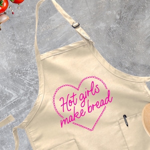 May include: A beige cotton apron with a pink heart design that says "Hot girls make bread".