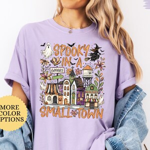 May include: An orchid-colored t-shirt featuring a graphic print with the text "SPOOKY IN A SMALL TOWN". The design includes illustrations of a ghost, a witch on a broomstick, a water tower, and various buildings decorated with autumn leaves.