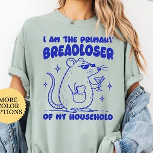 May include: A light green t-shirt featuring a blue graphic of a rat wearing sunglasses, holding a martini glass, and carrying a slice of bread in a pocket. The text reads I AM THE PRIMARY BREADLOSER OF MY HOUSEHOLD in bold blue lettering.