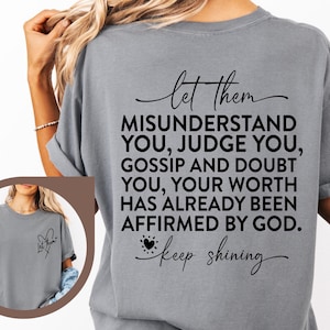 May include: Gray t-shirt with the text "let them MISUNDERSTAND YOU, JUDGE YOU, GOSSIP AND DOUBT YOU, YOUR WORTH HAS ALREADY BEEN AFFIRMED BY GOD. keep shining" printed on the back. The front of the shirt has a small heart with the text "let them" inside.