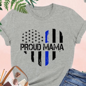 May include: Athletic heather gray t-shirt with a heart design. The heart features a black and blue striped design with stars and the words "PROUD MAMA" in black. The shirt is paired with blue jeans.