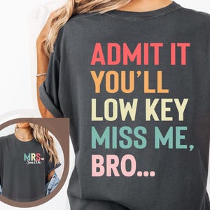 May include: A dark gray t-shirt with the text "ADMIT IT YOU'LL LOW KEY MISS ME, BRO..." printed in rainbow colors on the back. The front of the shirt has the text "MRS Smith" printed in rainbow colors.