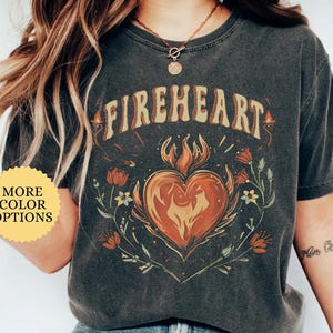 May include: Dark gray t-shirt with the word "FIREHEART" above a flaming heart design. The design includes floral elements and the text "MORE COLOR OPTIONS". The shirt is a Pepper color.
