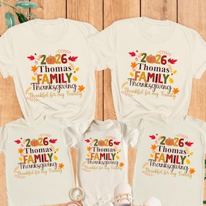 Op de afbeelding: Een collectie van natuurkleurige shirts en een babyrompertje met de tekst 2026 Thomas FAMILY Thanksgiving Thankful for my Family, versierd met oranje pompoenen en kleurrijke herfstbladeren in rood, geel en oranje.