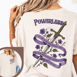 May include: Ivory-colored t-shirt with a graphic design on the back featuring a sword, purple flowers, and text that reads "POWERLESS" and a quote. The front of the shirt has a smaller floral design.