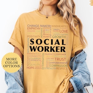 May include: Mustard-colored t-shirt with the words "SOCIAL WORKER" in large black letters. Surrounding the text are words like "EMPATHY," "TRUST," and "COMPASSION." The shirt also has a "MORE COLOR OPTIONS" button.