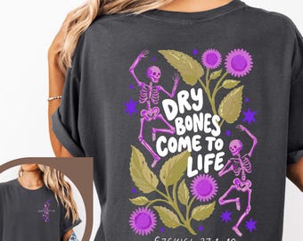 Puede incluir: Una camiseta gris carbón con un gráfico en la espalda de dos esqueletos rosas, hojas verdes y flores moradas. El texto dice "DRY BONES COME TO LIFE" y "EZEKIEL 37:1-10". Una versión más pequeña del gráfico del esqueleto aparece en el pecho izquierdo.
