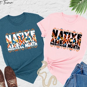 May include: Two t-shirts, one in heather deep teal and one in pink, with the text 'Native American Heritage Month' in colorful block letters with geometric patterns.