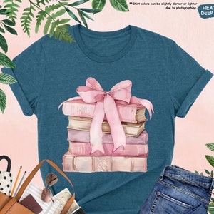 May include: A teal blue t-shirt with a watercolor illustration of a stack of books tied with a pink ribbon. The text "Shirt colors can be slightly darker or lighter due to photographing HEATHER DEEP TEAL" is printed on the shirt.