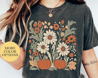 May include: A dark gray t-shirt featuring a floral design with daisies, orange flowers, and pumpkins. The text "PEPPER" is at the top right. A button on the left says "MORE COLOR OPTIONS".