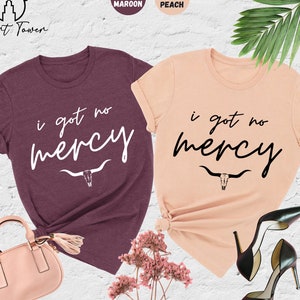 May include: Two t-shirts with the text "i got no mercy" and a skull and horns design. The shirt on the left is heather maroon and the shirt on the right is heather peach.