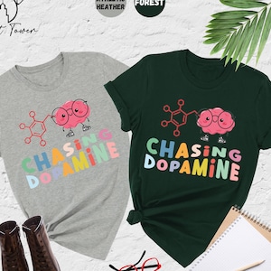 Pu&ograve; includere: Due t-shirt con una grafica di un cervello con gli occhiali e il testo "Chasing Dopamine" in colori arcobaleno. Le magliette sono in grigio melange atletico e verde bosco.
