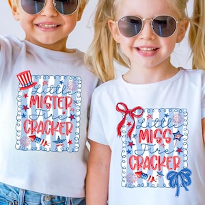 May include: White t-shirts with patriotic designs. One shirt says "Little Mister Fire Cracker" with a red, white, and blue design. The other shirt says "Little Miss Fire Cracker" with a similar design.