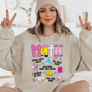 May include: A sand-colored crewneck sweatshirt featuring a graphic print with the word MATH in pink, yellow, and white patterns. Below, text reads Show your work, Do your best, Mistakes help you grow, and I believe in you, with math icons.