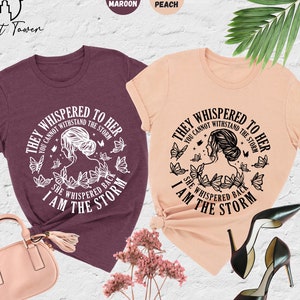 May include: Two t-shirts with a graphic design featuring a woman with butterflies and the text "They whispered to her you cannot withstand the storm she whispered back I am the storm". The t-shirts are in heather maroon and heather peach.