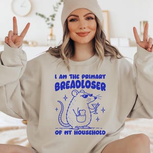 May include: A sand-colored crewneck sweatshirt featuring a blue graphic of a rat wearing sunglasses, holding a martini glass, and carrying a piece of toast in its pocket. The text reads: I AM THE PRIMARY BREADLOSER OF MY HOUSEHOLD.