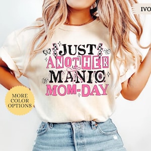 May include: Ivory-colored t-shirt with the text "JUST ANOTHER MANIC MOM-DAY" in black and pink. The shirt features decorative elements like butterflies and flowers. The words "MORE COLOR OPTIONS" are also visible.