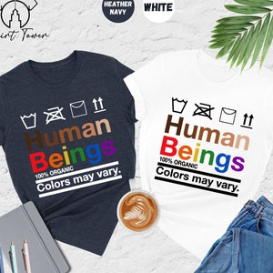 May include: Two t-shirts with the text "Human Beings" in rainbow colors and the words "100% Organic" and "Colors may vary." The t-shirts are in heather navy and white.