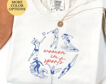 Puede incluir: Una camiseta de color marfil con un estampado gráfico azul de varias atletas y el texto rojo "women in sports". La camiseta tiene cuello redondo y una etiqueta de la marca Comfort Colors en el interior. Un collar con colgante dorado descansa encima.