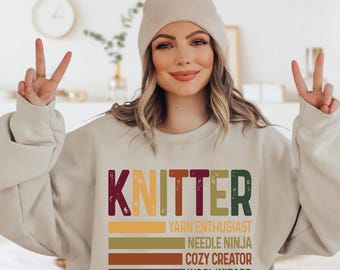 May include: A beige sweatshirt with the word "KNITTER" in a vintage style font. The text is layered with colorful stripes that read "Yarn Enthusiast", "Needle Ninja", "Cozy Creator", and "Wool Wizard".