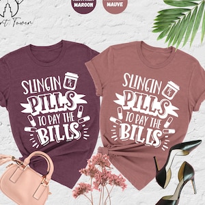 May include: Two heather colored t-shirts with the text "Slingin Pills to Pay the Bills" in white. The shirts are laid out on a white surface with a pair of black high heels and a pink purse.