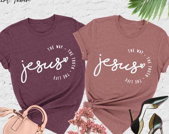 May include: Two heathered t-shirts, one maroon and one mauve, each with the word "Jesus" in a white script, encircled by "The Way, The Truth, The Life." The shirts are displayed with a purse, flowers, and high heels.