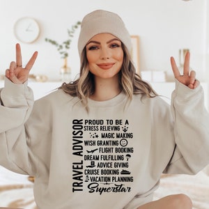 May include: A woman wearing a beige sweatshirt with the text "TRAVEL ADVISOR" and "PROUD TO BE A STRESS RELIEVING MAGIC MAKING WISH GRANTING FLIGHT BOOKING DREAM FULFILLING ADVICE GIVING CRUISE BOOKING VACATION PLANNING Superstar" printed on it.