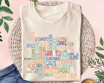 May include: A beige t-shirt with a word cloud design featuring the word "Thank You" in the center. The word cloud is made up of different languages, including "Merci", "Grazie", "Danke", "Obrigado", "Spasibo", and "Dyakuyu".