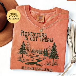 May include: A yam-colored t-shirt with the text "Adventure is Out There! But So Are Serial Killers" and a forest graphic. The shirt has a crew neck and short sleeves. A gold necklace with a pendant is draped across the shirt.