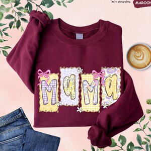 May include: Maroon sweatshirt with the word "MAMA" in a decorative font. The letters are in a variety of patterns, including gingham and floral designs. Pink bows and white flowers accent the letters. A cup of coffee and a pair of blue jeans are also in the image.