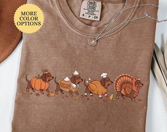 Puede incluir: Una camiseta color espresso con un gráfico de cuatro perros salchicha disfrazados para Acción de Gracias. Un perro lleva un disfraz de calabaza, otro es un pastel, uno es un pavo asado y otro es un pavo real. La camiseta tiene cuello redondo y etiqueta.