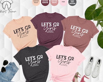 Peut inclure: Cinq t-shirts de différentes couleurs avec le texte "Let's Go Girls" et "Custom Destination" imprimé dessus. Les t-shirts sont en pêche chiné, bordeaux chiné, mauve chiné, rose et gris chiné foncé.