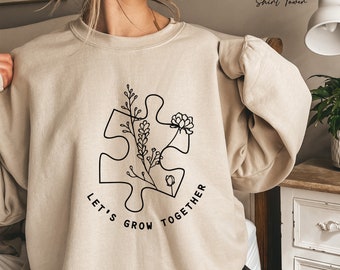 May include: A beige sweatshirt with a black outline of a puzzle piece with flowers growing from it. The text "Let's Grow Together" is written below the puzzle piece.