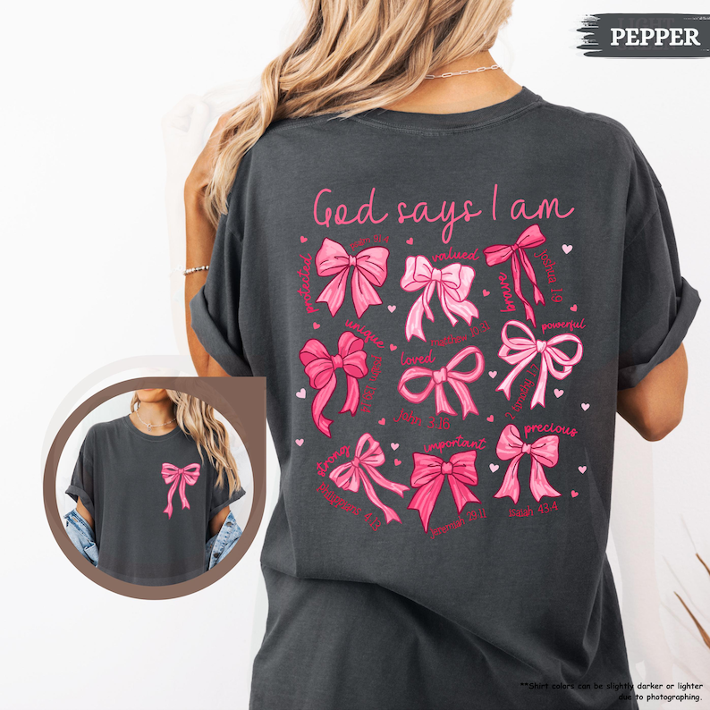 K&ouml;nnte beinhalten: Ein dunkelgraues T-Shirt mit einem rosa und wei&szlig;en Design, auf dem "Gott sagt, ich bin" steht, mit einer Reihe von rosa Schleifen und W&ouml;rtern wie "gesch&auml;tzt", "geliebt", "stark", "wichtig" und "kostbar".