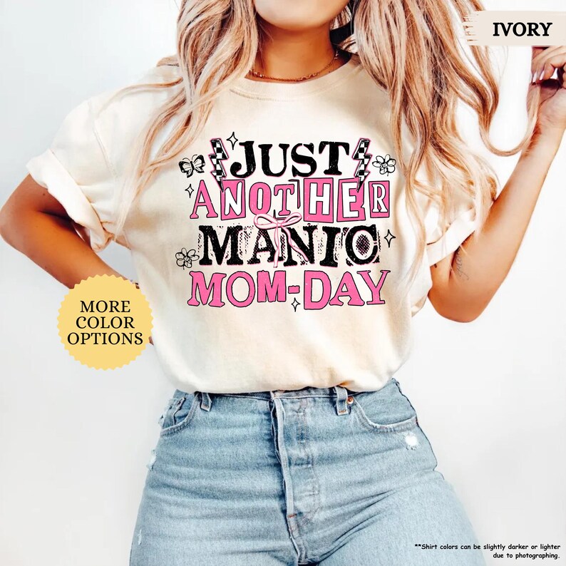 May include: Ivory-colored t-shirt with the text "JUST ANOTHER MANIC MOM-DAY" in black and pink. The shirt features decorative elements like butterflies and flowers. The words "MORE COLOR OPTIONS" are also visible.