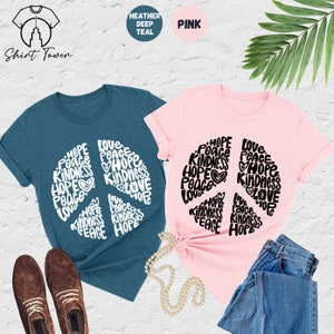 May include: Two t-shirts with a peace sign design. The peace sign is made up of the words "Hope", "Peace", "Love", and "Kindness". One shirt is heather deep teal and the other is pink.
