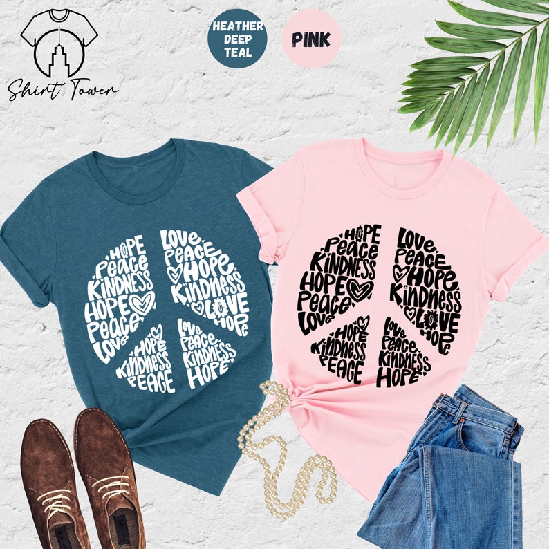 May include: Two t-shirts with a peace sign design. The peace sign is made up of the words "Hope", "Peace", "Love", and "Kindness". One shirt is heather deep teal and the other is pink.