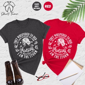 May include: Two t-shirts with a white graphic design featuring a woman with butterflies and the text "They whispered to her you cannot withstand the storm she whispered back I am the storm". The t-shirts are in dark heather grey and red.
