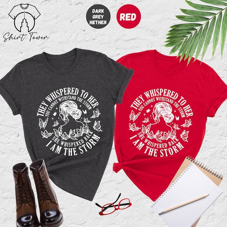 May include: Two t-shirts with a white graphic design featuring a woman with butterflies and the text "They whispered to her you cannot withstand the storm she whispered back I am the storm". The t-shirts are in dark heather grey and red.
