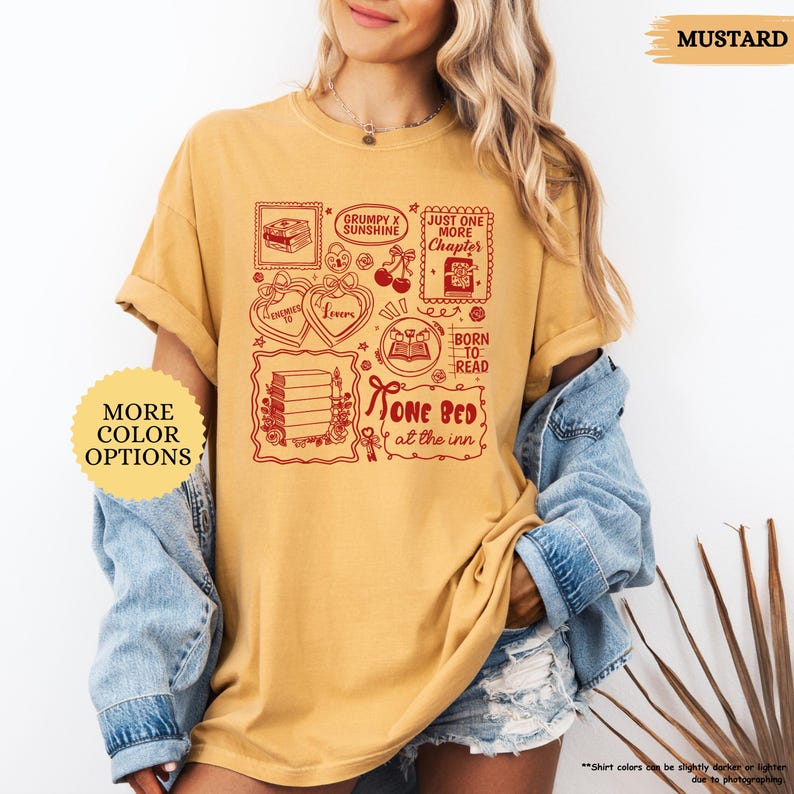 May include: Mustard-colored t-shirt with a red graphic featuring book-themed illustrations and text. The design includes phrases like "Grumpy x Sunshine," "Just One More Chapter," and "One Bed at the Inn." The shirt has a relaxed fit.