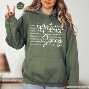 May include: Military green hoodie with a front pocket and a drawstring hood. The hoodie features the words "Mentally Spicy" in a white script font, along with a list of descriptive words. The hoodie is made of a soft material.