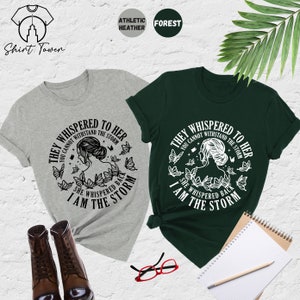 May include: Two t-shirts with a graphic design featuring a woman with butterflies and the text "They whispered to her you cannot withstand the storm she whispered back I am the storm". The t-shirts are in athletic heather grey and forest green.
