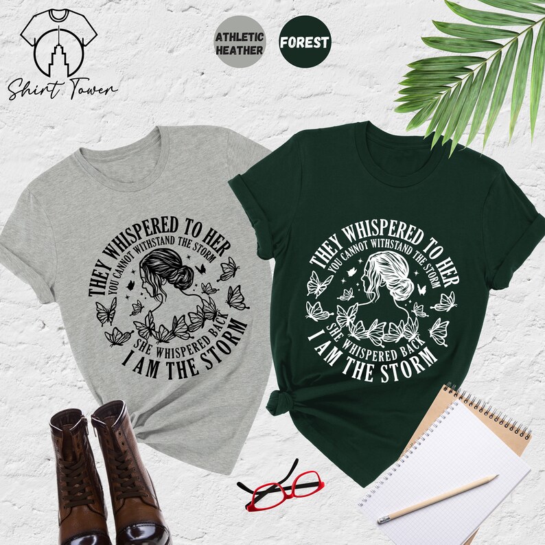 May include: Two t-shirts with a graphic design featuring a woman with butterflies and the text "They whispered to her you cannot withstand the storm she whispered back I am the storm". The t-shirts are in athletic heather grey and forest green.